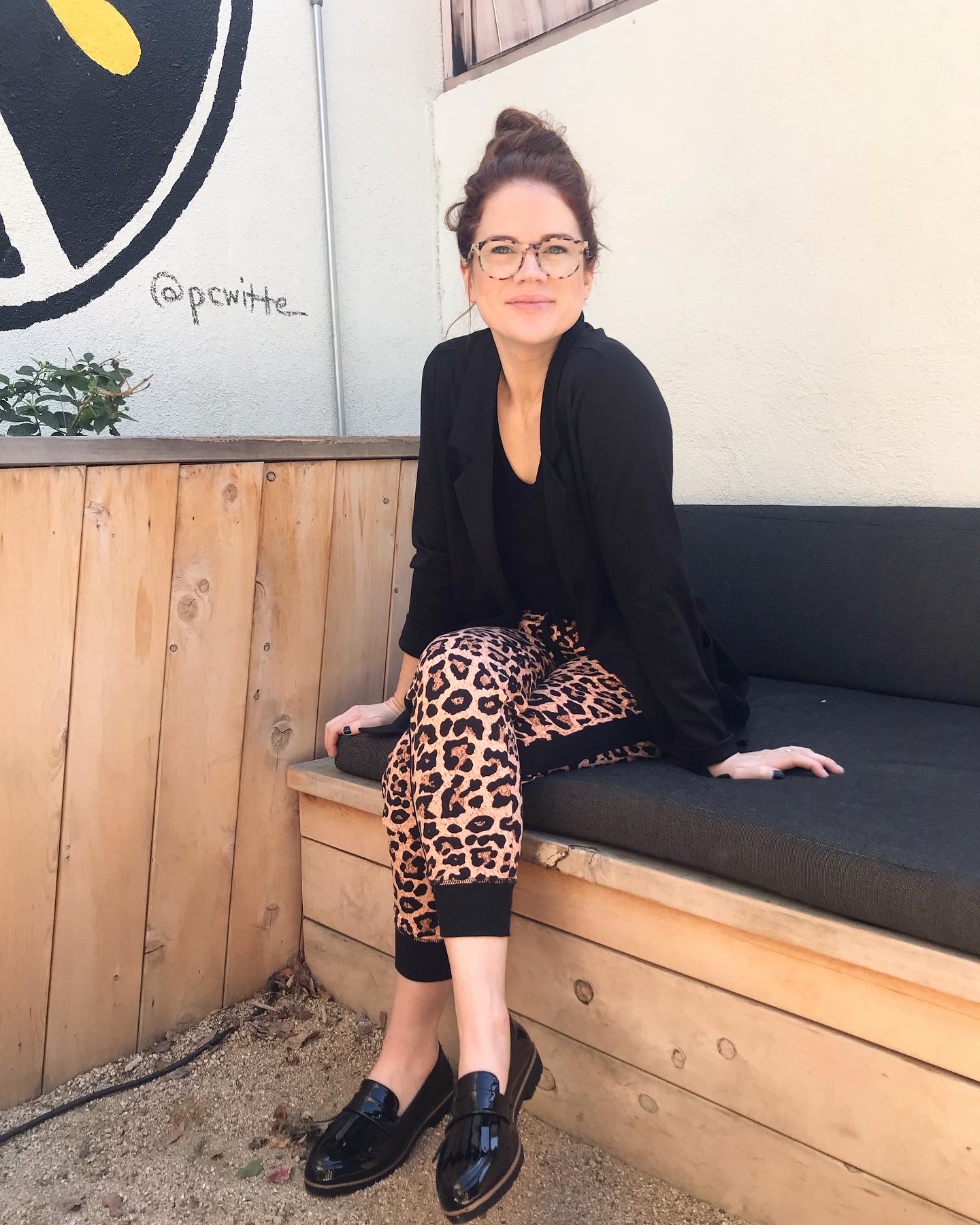 Katie Dunlavy, Lularoe boutique owner, of The Pearl Pages sitting on a black and wood bench wearing a black shirt and blazer with animal print joggers and loafers, with glasses and hair in a top knot